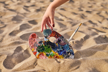 Hand adjusts colorful artist palette and brush on sunlit sandy beach, symbolizing creativity and...
