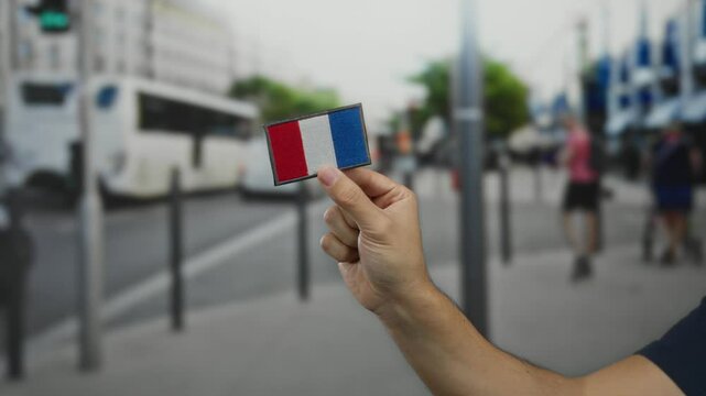 Man holding embroidered france flag patch on city street background showcasing patriotism and outdoor urban scene familiar in european towns.