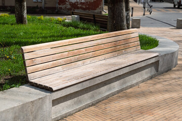 Wooden bench with a modern minimalist design stands on a curved concrete base in a green city park, illuminated by soft sunlight.