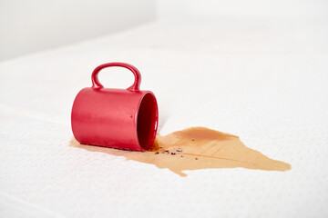 Red mug lies on white surface, spilling liquid and forming brown stain. Bright lighting emphasizes...