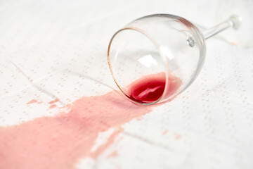 Red wine stain spreads across white textured fabric, highlighting contrast. Empty glass lies on...