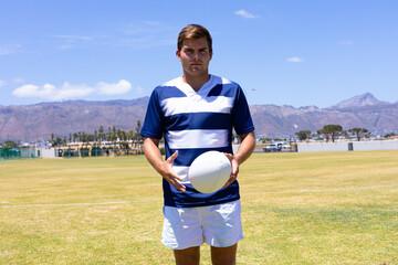 Man wearing striped sports jersey holding white rugby ball on field under sunny sky with mountains