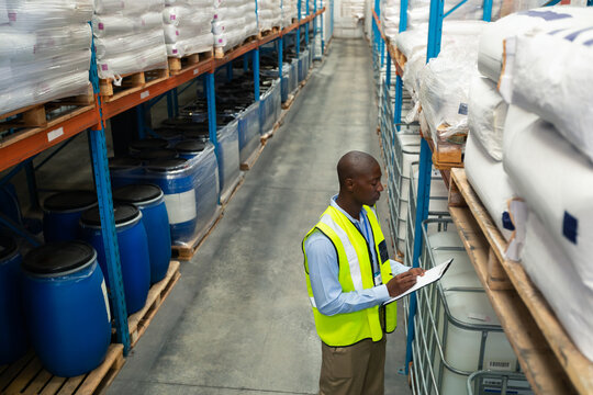 Warehouse worker inspecting pallets stacked with white sacks and blue barrels in warehouse aisle