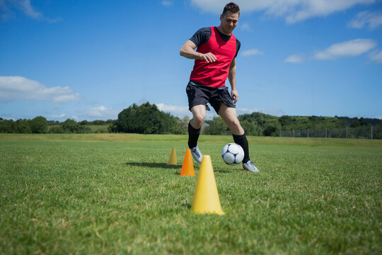 Male athlete dribbling soccer ball around training cones on grass field wearing red bib and cleats
