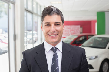 Fototapeta premium Middle-aged man wearing suit and striped tie standing by display cars in showroom by glass windows