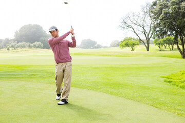 Asian man standing on putting green holding golf iron during sunny day on tree-lined fairway