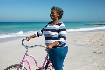 Naklejka premium Senior African American woman standing on sandy beach holding pink cruiser bicycle, copy space