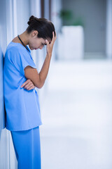 Nurse leaning against wall in bright hospital corridor, holding forehead and wearing stethoscope