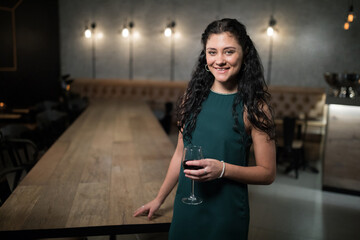 Woman leaning against table at bar in green dress holding glass of red wine, copy space