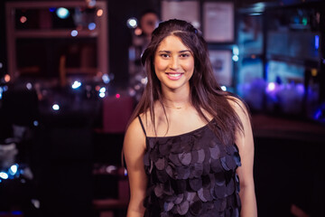 Woman in her twenties wearing sequined top posing in dimly lit bar with illuminated bottles