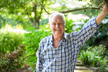 Senior man leaning on tree branch while smiling in backyard garden with stepping-stone path