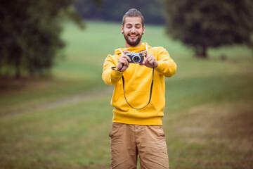 Man standing in grassy field wearing yellow hoodie and brown pants holding vintage camera