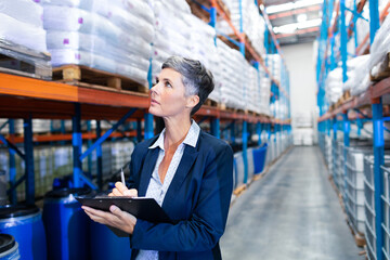 Woman wearing navy blazer with clipboard inspecting pallet racks and barrels in warehouse aisle