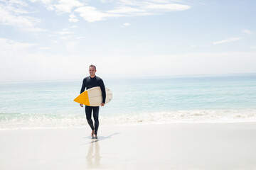 Male surfer walking barefoot on sandy beach by turquoise sea carrying surfboard with yellow nose
