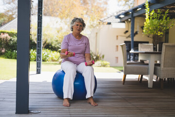 Senior woman sitting on blue exercise ball on deck holding pink dumbbells by wicker dining set
