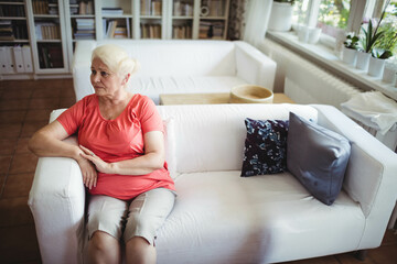 Obraz premium Senior woman sitting on sofa in living room wearing coral top beside built-in bookshelf