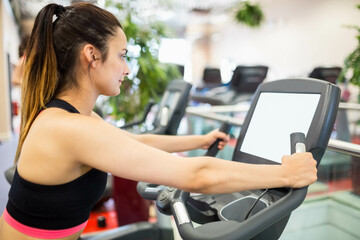 Fototapeta premium Female fitness enthusiast gripping elliptical handles monitoring digital console at gym, copy space