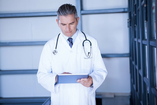 Male doctor wearing lab coat with stethoscope reviewing tablet in corridor with shelving units