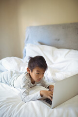 Chinese boy wearing striped pajamas lying on stomach on bed at home using silver laptop