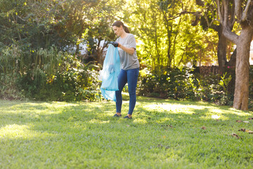 Woman bending, picking up debris in sunlit backyard wearing black gloves, holding blue plastic bag