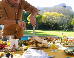 Middle-aged man carving roast turkey with knife and fork on patio table by pool, copy space