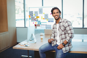 Indian man holding eyeglasses and smiling while sitting on office desk with laptop and coffee cup