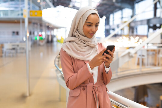 Woman wearing trench coat and headscarf leaning on railing inside terminal checking smartphone