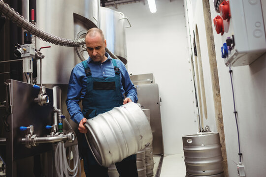 Male brewer wearing work overalls handling metal beer keg in brewery with fermentation tanks - Powered by Adobe