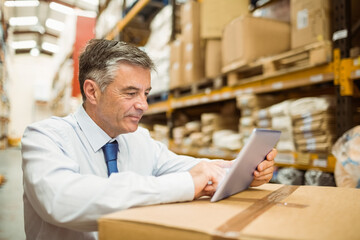 Senior white male manager in business attire standing in warehouse aisle examining tablet on box