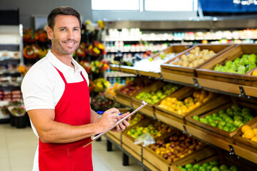 Male in red apron inspecting fruits at wooden produce bins holding clipboard and pen, copy space