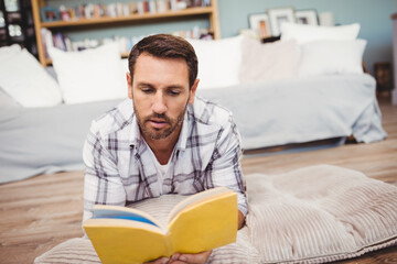 Man lying on cushioned mat on wooden floor reading yellow hardcover book near daybed and bookshelf