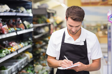 male grocery employee in apron writing notes on notepad in produce aisle, cold shelves, copy space