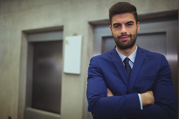 Man in navy suit standing with arms crossed in office lobby by elevator doors, copy space