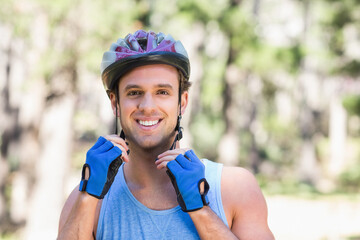 Man adjusting bicycle helmet straps while standing in wooded park wearing blue cycling gloves