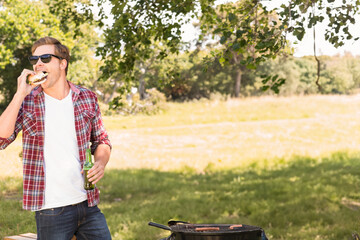 Charcoal grill cooking sausages over hot coals in sunlit grassy park, burger and beer nearby