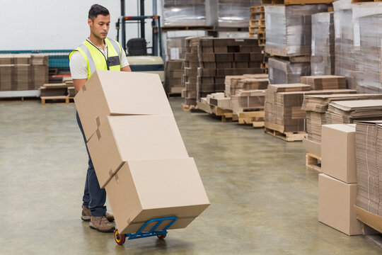 Hispanic male in safety vest pushing blue hand truck with cardboard boxes across warehouse floor - Powered by Adobe