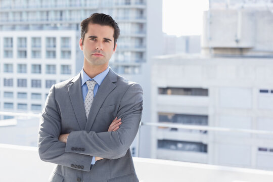 Businessman in suit standing with arms crossed on rooftop terrace overlooking cityscape and railing - Powered by Adobe