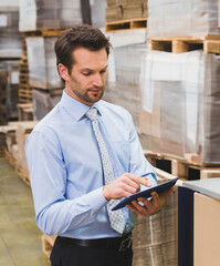 Male supervisor in shirt and tie reviewing inventory on tablet at workstation with boxes on pallets