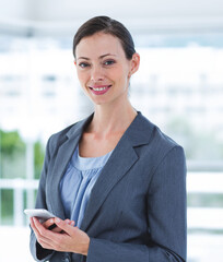 Woman wearing grey blazer and light blue blouse, holding smartphone while smiling in office lobby