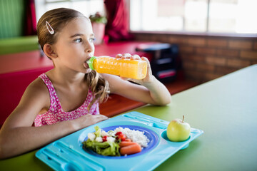 Girl sipping orange juice from bottle in cafeteria, holding blue tray with rice veggies and apple © wavebreak3