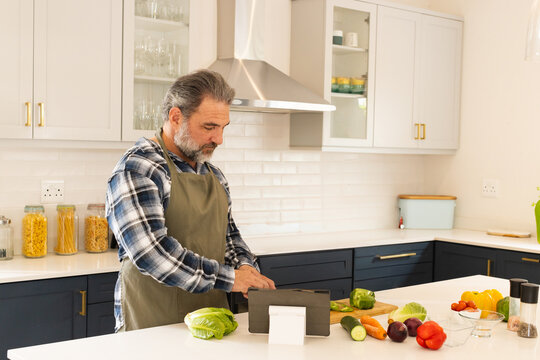 Senior man wearing green apron chopping vegetables on wooden board in kitchen while using tablet