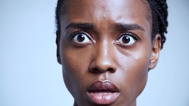 Shocked african american woman with wide eyes and open mouth displaying intense fear and surprise against pale blue backdrop