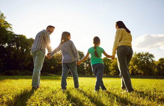 Back view of father, mother, daughter and son holding hands together and standing in fiend on green grass, admiring sunshine happy to live in this worls and enjoying life. Family time