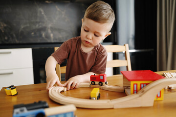 A young boy plays with a wooden toy train set at home, carefully connecting the tracks. The child is focused and engaged in creative and educational play at the kitchen table. © Aigul Sabirova
