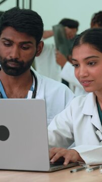 Vertical video. Diverse indian medical professionals reviewing patient data on laptop, showcasing collaborative expertise in modern hospital setting with medical staff in background