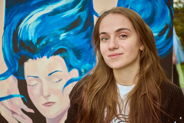 Young adult caucasian female with long brown hair smiling beside colorful mural. Artwork behind her...