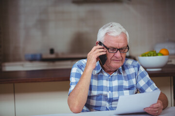 Senior male calling on smartphone while reading document at kitchen table with fruit bowl