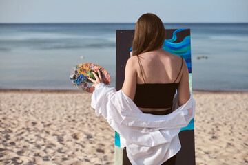 Young female painter stands by sea, absorbed in creating vibrant artwork on canvas. Beach setting...