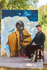 Young adult female with short hair sits beside large outdoor painting depicting back view of woman...