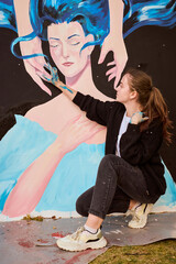 Young woman with brown hair painting large mural outdoors. Focused expression, wearing black sweater, white shirt. Blue tones dominate artwork depicting abstract face